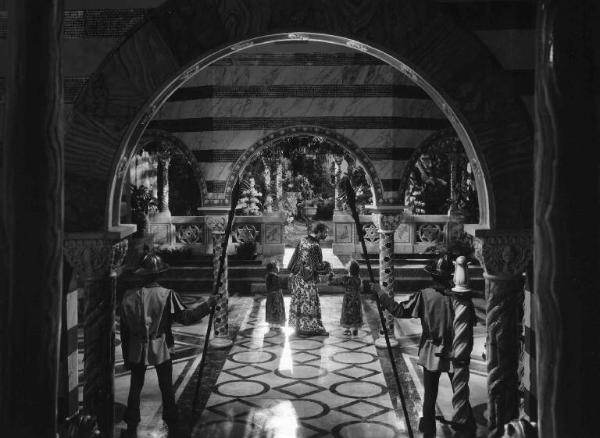 Set del film "La corona di ferro" - Regia Alessandro Blasetti - 1940 - Un attore adulto non identificato da la mano a due attori bambini non identificati all'interno di una sala sorvegliata da soldati.