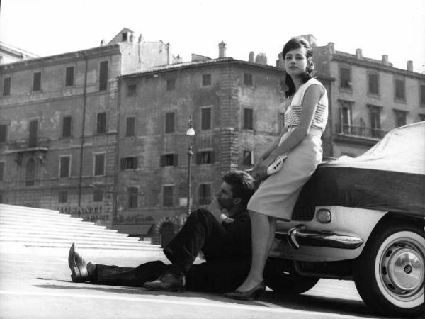 Fotografia del film "La cuccagna" - Regia Luciano Salce 1962 - L'attore Luigi Tenco e l'attrice Donatella Turri appoggiati ad un'automobile in una piazza.