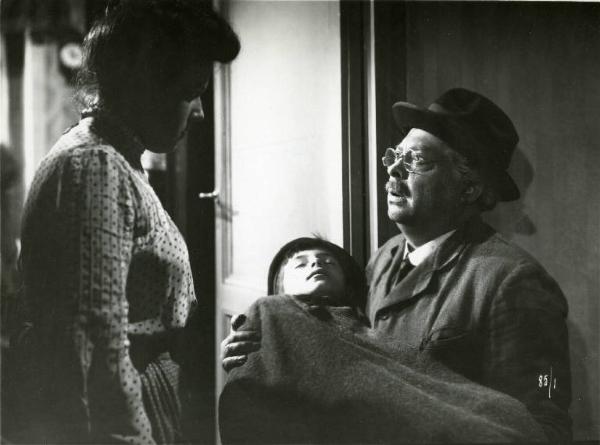 Scena del film "Il delitto di Giovanni Episcopo" - Regia Alberto Lattuada, 1947 - Aldo Fabrizi tiene in braccio il figlio, nella vita e nel film, Amedeo privo di sensi mentre dialoga con una donna, in piedi di fronte a lui.