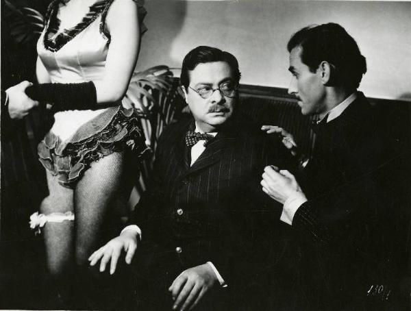 Scena del film "Il delitto di Giovanni Episcopo" - Regia Alberto Lattuada, 1947 - Aldo Fabrizi, seduto su un divanetto, guarda un uomo seduto accanto a lui. Alla sua sinistra una ballerina inquadrata dalle ginocchia al collo.