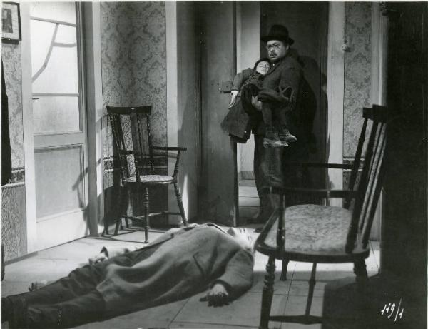 Scena del film "Il delitto di Giovanni Episcopo" - Regia Alberto Lattuada, 1947 - In primo piano giace supino il cadavere di Roldano Lupi. Aldo Fabrizi, con in braccio il figlio, in piedi accanto alla porta, lo guarda corrucciato.