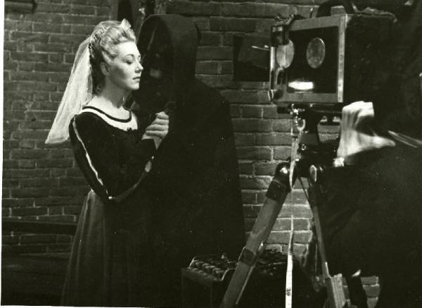 Sul set del film "Dente per dente" - Regia Marco Elter, 1943 - Piano americano di Nelly Corradi e Alfredo Varelli. L'attore travestito da frate, indossa una maschera e tiene le mani di lei. In primo piano la cinepresa.