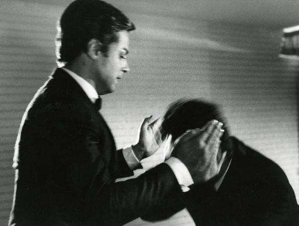 Scena del film "Diabolik" - Regia Mario Bava, 1968 - Mezza figura di John Phillip Law con le mani parallele all'altezza delle orecchie di un uomo, in movimento, chinato dinanzi a lui.