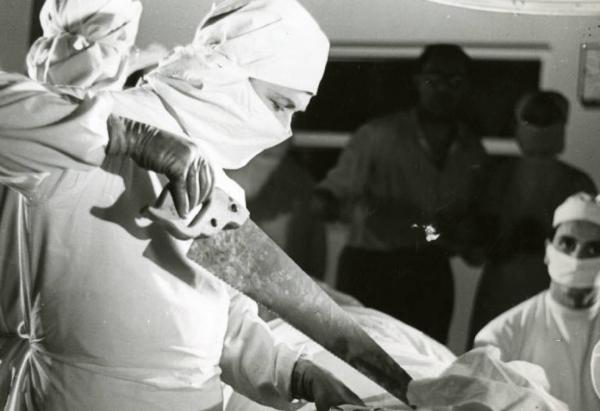 Sul set del film "Diagnosi" - Regia Ferruccio Cerio, 1942 - Un attore, nei panni di un chirurgo, sta operando con un sega. Sullo sfondo, in penombra, Ferruccio Cerio.