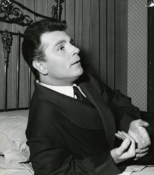 Scena del film "Difendo il mio amore" - Regia Giulio Macchi, 1956 - Mezza figura di profilo di Gabriele Ferzetti, in abiti eleganti, appoggiato a un letto con il braccio destro.