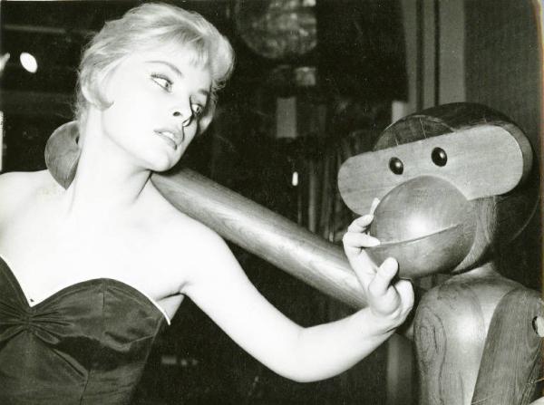 Scena del film "La donna del giorno" - Regia Francesco Maselli, 1957 - Mezza figura di Virna Lisi. L'attrice ha una mano sul muso di una statua di legno a forma di scimmia, che con una zampa a forma di uncino, le cinge il collo da dietro.