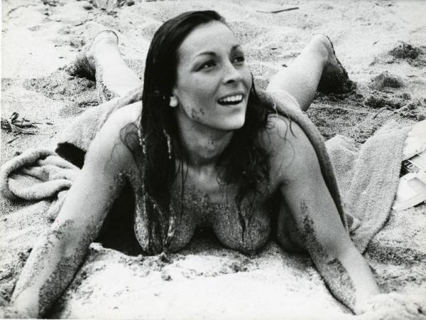 Scena del film "Una donna... e una donna" - Regia Luigi Petrini, 1969 - Figura intera. Susanna Levi prona sulla spiaggia, solleva testa e spalle e mostra il seno nudo, coperto di sabbia. Un asciugamano le copre schiena e glutei.