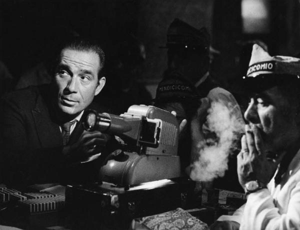 Scena del film "La donna scimmia" - Regia Marco Ferreri, 1964 - Mezza figura di Ugo Tognazzi mentre sistema un proiettore di diapositive. A destra, un uomo fuma una sigaretta.
