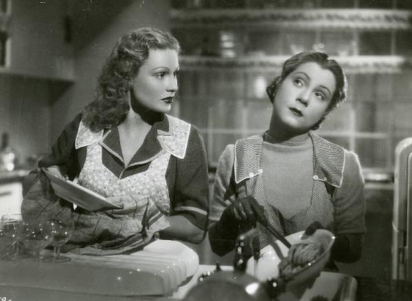Scena del film "Dopo divorzieremo" - Regia Nunzio Malasomma, 1940 - Mezza figura di Vivi Gioi mentre asciuga un piatto e guarda Lilia Silvi, al suo fianco, che lava un'insalatiera con una spazzola lavapiatti.