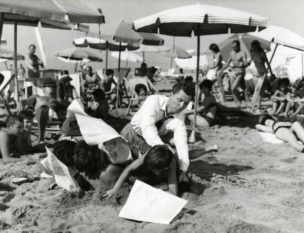 Scena del film "Una domenica d'estate" - Regia Giulio Petroni, 1962 - In spiaggia una donna in una buca nella sabbia è travolta dalle pagine di un giornale e da due bambini. Eddie Bracken davanti a loro cerca di afferrarne uno. Sullo sfondo bagnanti.