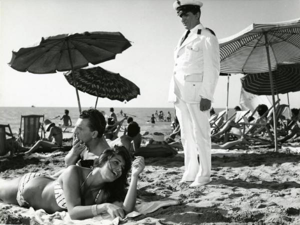 Scena del film "Una domenica d'estate" - Regia Giulio Petroni, 1962 - In spiaggia Eddie Bracken, seduto, e Francoise Fabian sdraiata, prona, su un asciugamano. In piedi, dietro di loro, un vigile urbano li guarda. Sullo sfondo, sdraio e bagnanti.