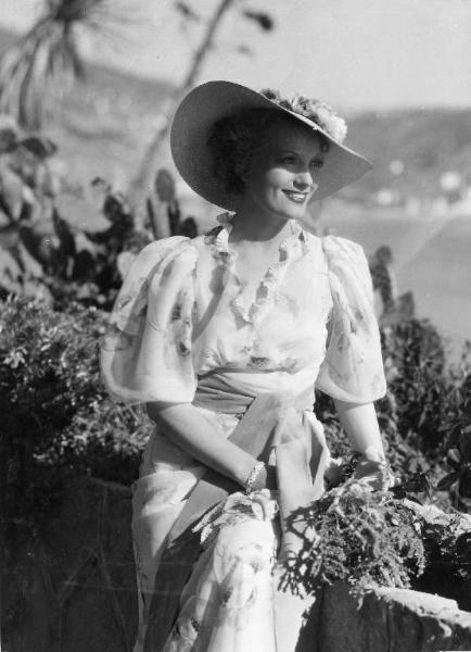 Scena del film "Una donna fra due mondi" - Regia Goffredo Alessandrini, 1936 - Piano americano di Isa Miranda seduta su un muretto. L'attrice, tiene un mazzo di fiori di campo in mano.