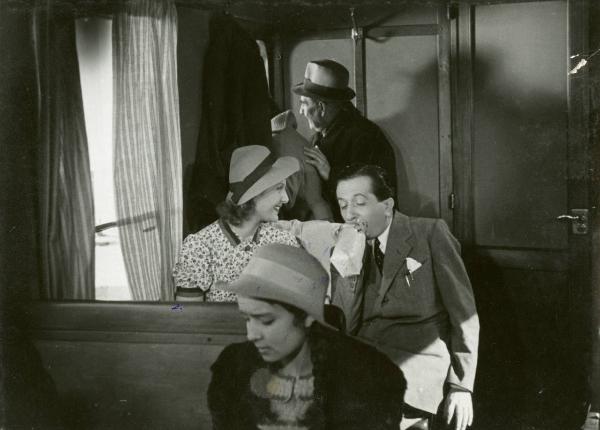 Scena del film "Due milioni per un sorriso" - Regia Carlo Borghesio, Mario Soldati, 1939 - Enrico Viarisio soffia in un sacchetto di carta mentre Elsa De Giorgi lo osserva divertita. Entrambi sono seduti in treno, con attorno attori non identificati.