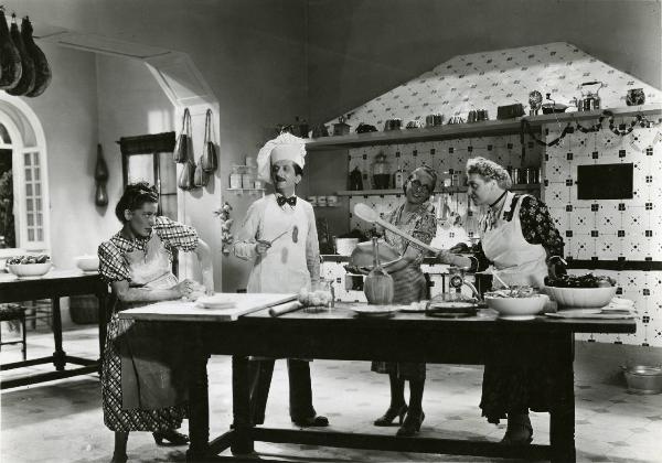Scena del film "Duetto vagabondo" - Regia Guglielmo Giannini, 1938 - Al centro, Enrico Viarisio con cappello da cuoco, Pina Renzi, a destra con un cucchiaio di legno in mano, e attrici non identificate cucinano.