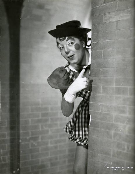 Scena del film "È bello qualche volta andare a piedi" - Regia Michele Galdieri, 1941 - Un'attrice non identificata, vestita e truccata da clown, appare da dietro una parete e indica con la mano destra.