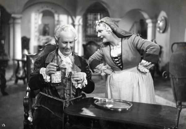 Scena del film "Elisir d'amore" - Regia Amleto Palermi, 1941 - Olinto Cristina, seduto, regge nella mano sinistra un bicchiere e nella destra una bottiglia con l'etichetta "Elisir di Lunga Vita". A sinistra, un'attrice non identificata.