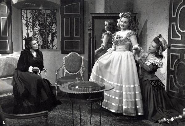 Scena del film "Elisir d'amore" - Regia Amleto Palermi, 1941- A sinistra, Pina Renzi, seduta su un divano, osserva un'attrice non identificata che aiuta Gemma D'Alba, a destra, a vestirsi.