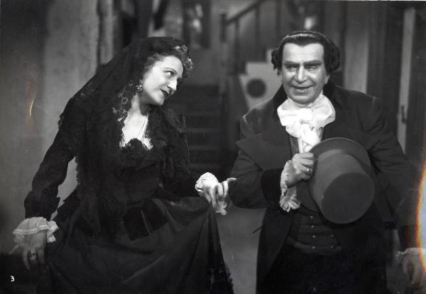 Scena del film "Elisir d'amore" - Regia Amleto Palermi, 1941- A sinistra, Pina Renzi, sollevando la gonna con entrambe le mani, e a destra, Armando Falconi, con un cappello in mano, si guardano sorridenti.