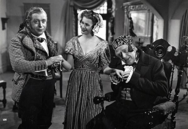 Scena del film "Elisir d'amore" - Regia Amleto Palermi, 1941- A sinistra, un attore non identificato e, a destra, Armando Falconi guardano dritto davanti a loro, mentre tengono per mano Gemma D'Alba, al centro, che guarda sorridente verso sinistra.