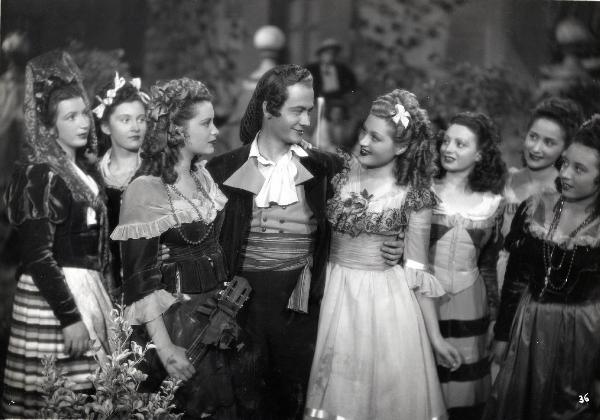 Scena del film "Elisir d'amore" - Regia Amleto Palermi, 1941 - Roberto Villa, al centro, stringe a sé Jone Salinas, a sinistra, mentre guarda Livia Minelli, a destra. Intorno, attrici non identificate osservano la scena.
