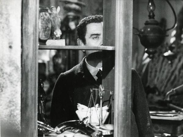 Scena del film "Er più storia d'amore e di coltello" - Regia Sergio Corbucci, 1971 - Vittorio Caprioli, nascosto dietro a una vetrinetta contenente una pistola, osserva dritto davanti a sé.