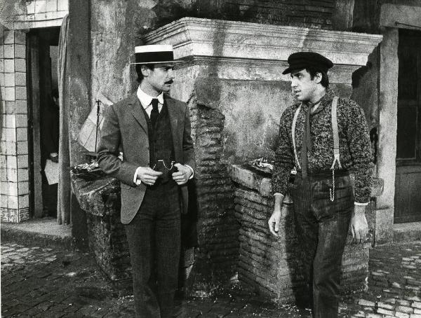 Scena del film "Er più storia d'amore e di coltello" - Regia Sergio Corbucci, 1971 - Gianni Macchia, a sinistra, con la mano sinistra in tasca, e Adriano Celentano, a destra, si guardano.