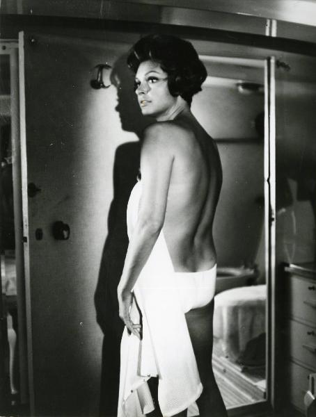 Scena del film "L'estate" - Regia Paolo Spinola, 1966 - Mezza figura di un'attrice non identificata, di spalle, con la schiena nuda, che si copre con un asciugamano e rivolge lo sguardo a sinistra.
