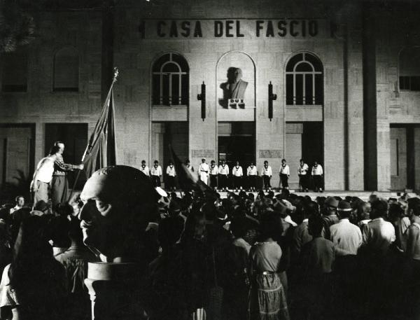Scena del film "Estate violenta" - Regia Valerio Zurlini, 1959 - Attori non identificati osservano in direzione di un edificio recante la scritta "CASA DEL FASCIO". All'ingresso è schierata una fila di attori non identificasti che guardano la folla.