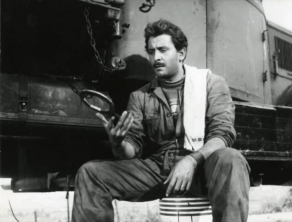 Scena del film "Esterina" - Regia Carlo Lizzani, 1959 - Davanti a un treno, seduto su un barile, Domenico Modugno rivolge lo sguardo verso sinistra mentre gioca con un oggetto non identificato.