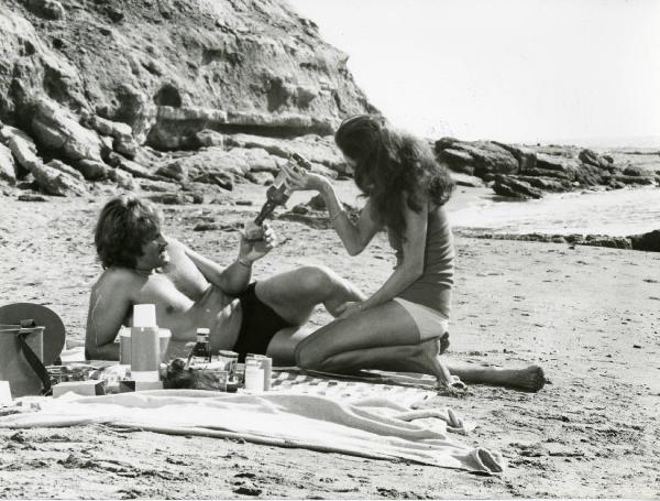 Scena del film "L'etrusco uccide ancora" - Regia Armando Crispino, 1972 - Su una spiaggia, con teli e cibo da pic nic: Samantha Eggar, inginocchiata, versa da bere ad Alex Cord, disteso con un bicchiere di vetro in mano.