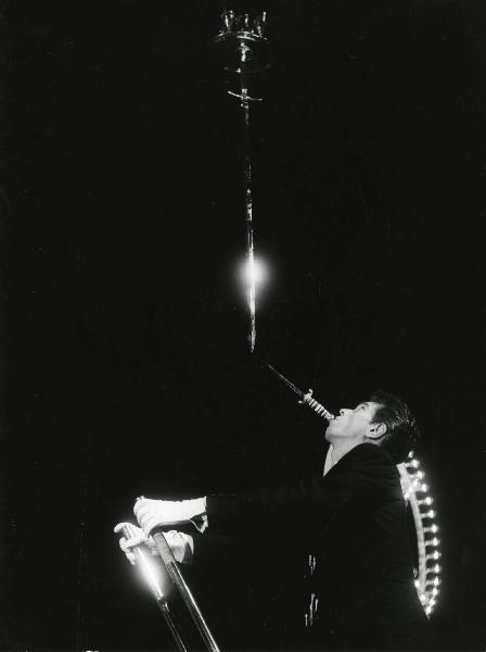 Scena del film "Europa di notte" - Regia Alessandro Blasetti, 1959 - Un attore non identificato, si regge a una scala con le mani e sostiene con la bocca un pugnale e una spada sulla quale è appoggiato un vassoio con alcuni bicchieri.