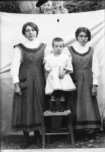 Val Brembana. Ritratto di due ragazze con bambino al centro in piedi su una sedia