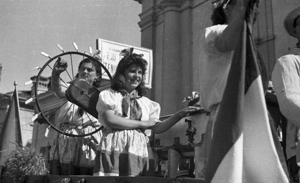 Garlasco - Piazza della Repubblica (già Vittorio Emanuele II) - Festa delle mondine - Carro con due giovani donne in abiti tradizionali, un cartellone parzialmente occultato - In primo piano due persone con bandiera italiana parzialmente visibili