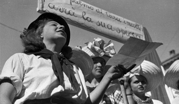 Garlasco - Festa delle mondine - Giovani donne in abiti tradizionali, di cui una tiene un foglio in mano - Cartello con scritta "Ogni palmo di terra incolto avrà la sua spiga"