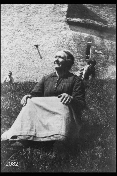 Giovanna Canella. Donna anziana seduta sul prato davanti a cascina.