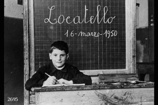 Scolaro di Locatello, in classe. - Posa in interno alle spalle dello scolaro una lavagna colla scritta: "Locatello / 16 - marzo - 1950".
