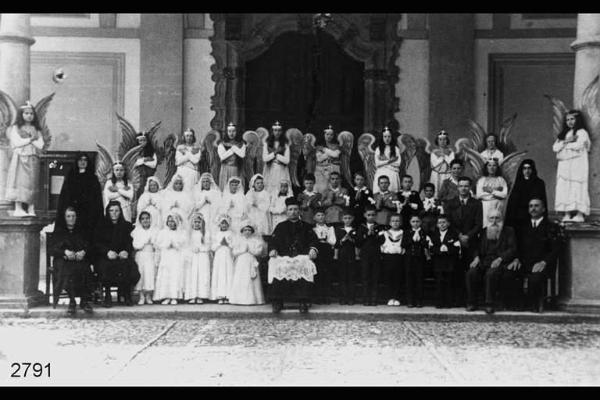 Ritratto di gruppo in occasione della prima comunione. Don Angelo Mazzoleni tra un gruppo di bambini e bambine in abito da cerimonia, e altri vestiti da angeli, sul sagrato di una chiesa.