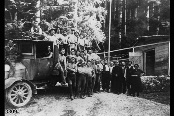 Lavoratori emigranti di Capizzone e Berbenno posano in un bosco con camion e capanno sullo sfondo.
