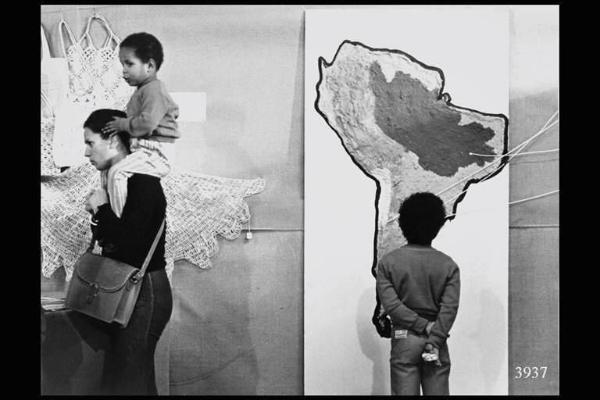 Milano. Parco Sempione. Festa dell'Unità: ragazzo osserva una carta geografica dell'America del Sud; a sinistra, donna con bambino sulle spalle.