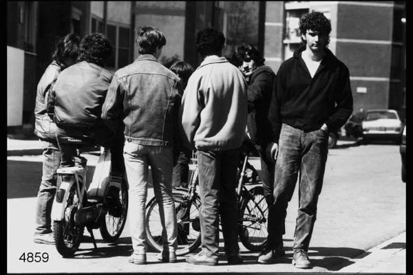 Quarto Oggiaro. Gruppo di giovani, in parte di spalle. Due sono seduti, l'uno su di un motorino, l'altro su di una bicicletta.