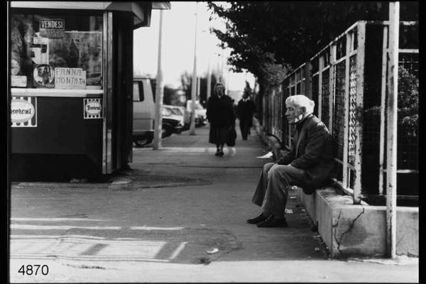 Quarto Oggiaro. Marciapiede di una via del quartiere.Sul lato sinistro un chiosco, sul lato destro un anziano seduto sul bordo di una cancellata.