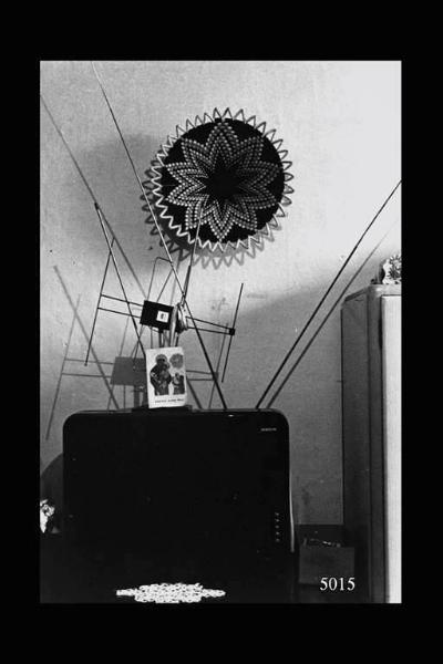 Interno di abitazione eritrea. Televisore, alla sommità immaginetta religiosa sorretta dall'antenna dell'apparecchio, sullo sfondo decorazione tradizionale a muro.