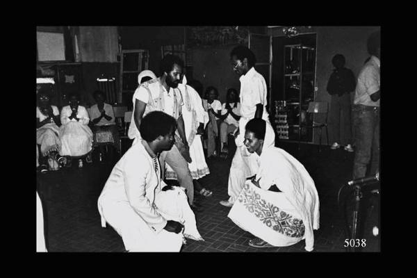Eritrei a Milano. Gruppo di eritrei danza al centro del locale. In primo piano coppia inchinata in una fase della danza. Sullo sfondo donne sedute accompagnano la danza battendo le mani.