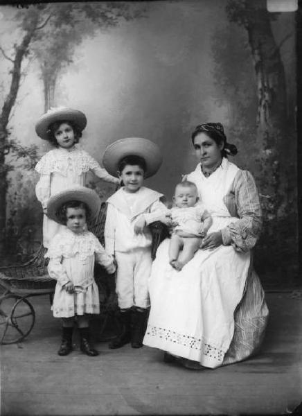 Balia di Tonia coi suoi bambini - Posa in studio. La balia con il consueto costume e grembiule bianco i tre bambini in tenuta elegante con grandi cappelli di paglia.