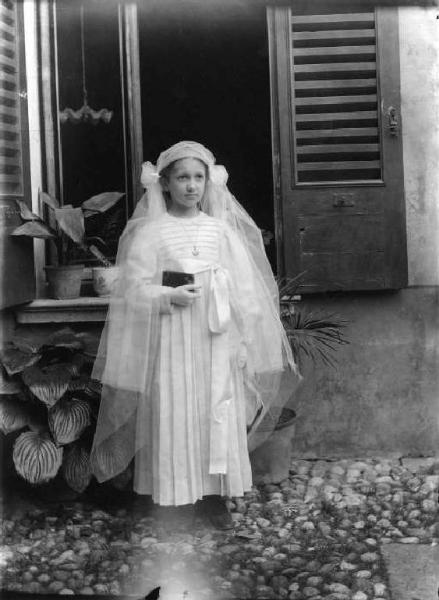 Annunciata alla Prima Comunione. Bambina in posa, all'esterno di una casa, in abito da prima comunione.