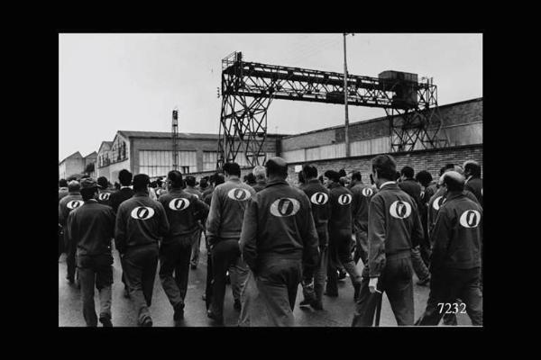 Innocenti Leyland. Gruppo di lavoratori in tuta da lavoro, di spalle, all'interno della fabbrica. Probabilmente in partenza per un corteo sindacale.