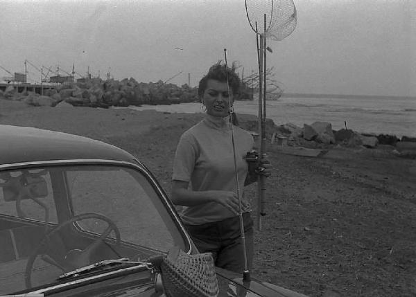 Località non identificata. Sofia Loren, vicino ad un'automobile, sulla spiaggia. L'attrice tiene in mano una canna e un retino da pesca
