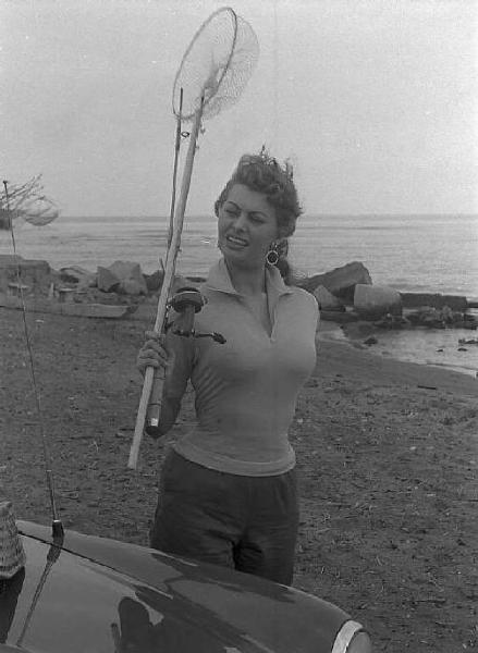 Località non identificata. Sofia Loren, vicino ad un'automobile, sulla spiaggia. L'attrice tiene in mano un canna e un retino da pesca