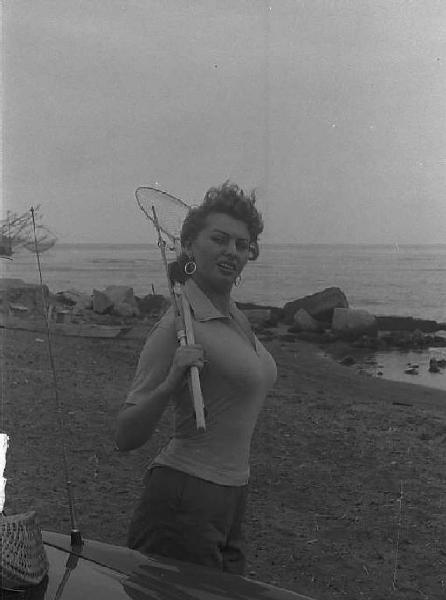 Località non identificata. Sofia Loren, vicino ad un'automobile, sulla spiaggia. L'attrice tiene in mano una canna e un retino da pesca