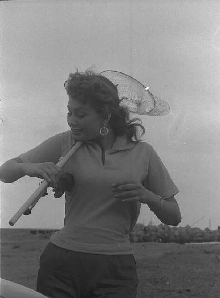 Località non identificata. Sofia Loren sulla spiaggia. L'attrice tiene in mano una canna e un retino da pesca